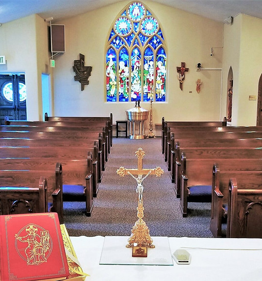 A Catholic church with brown carpets, wooden pews, and a gold crucifix taken from the viewpoint of the priest facing the congregation